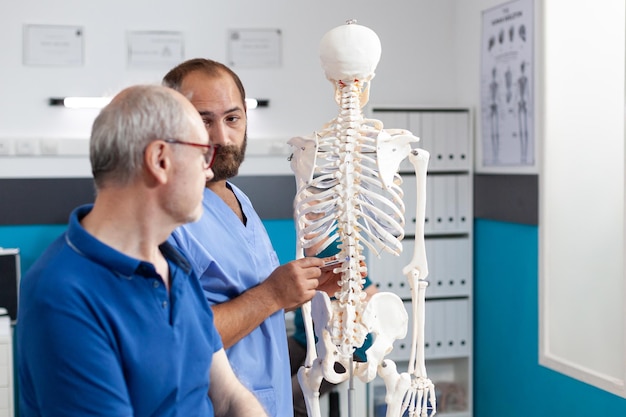 Ortopedista explicando la médula espinal en el esqueleto humano al paciente jubilado en la oficina de fisioterapia. Quiropráctico haciendo presentación de huesos y anatomía de la columna a anciano por osteopatía