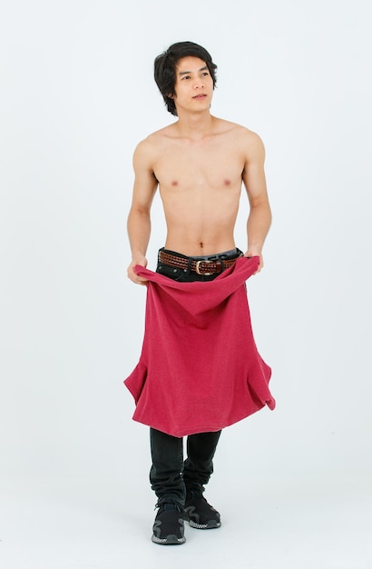 Ortrait isolierte Ausschnitt-Ganzkörper-Studioaufnahme eines asiatischen jungen, gutaussehenden, selbstbewussten, schlanken, hemdlosen Teenager-Männchenmodells, das mit einem roten T-Shirt in den Händen steht und einen muskulösen Oberkörper auf weißem Hintergrund zeigt