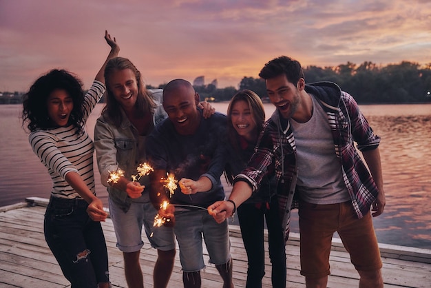 Os amigos fazem-se felizes. Grupo de jovens em roupas casuais sorrindo e segurando faíscas