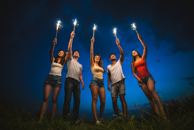 Os amigos sorridentes seguram bastões de fogos de artifício. noite noite