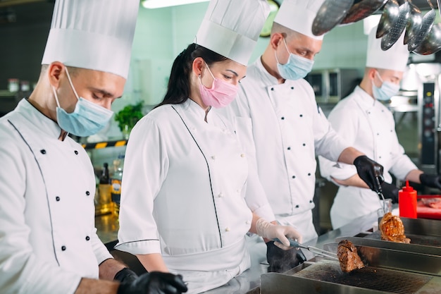 Os chefs em luvas e máscaras protetoras preparam os alimentos na cozinha de um restaurante ou hotel.