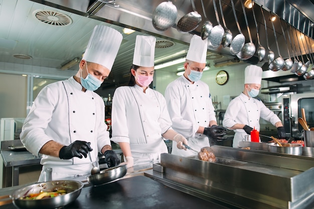 Os chefs em luvas e máscaras protetoras preparam os alimentos na cozinha de um restaurante ou hotel.