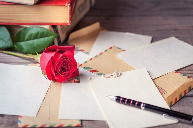 Os envelopes do vintage, a rosa do vermelho e as folhas de papel dispersaram na tabela de madeira para escrever letras românticas.