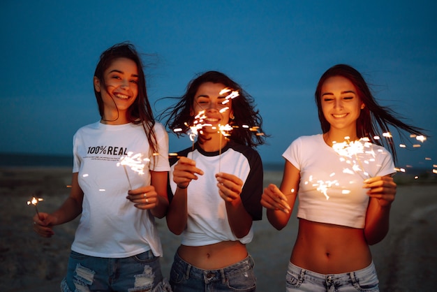 Os estrelinhas nas mãos de jovens na praia. três meninas curtindo a festa na praia com acendedor. férias de verão, férias, relaxar e conceito de estilo de vida.