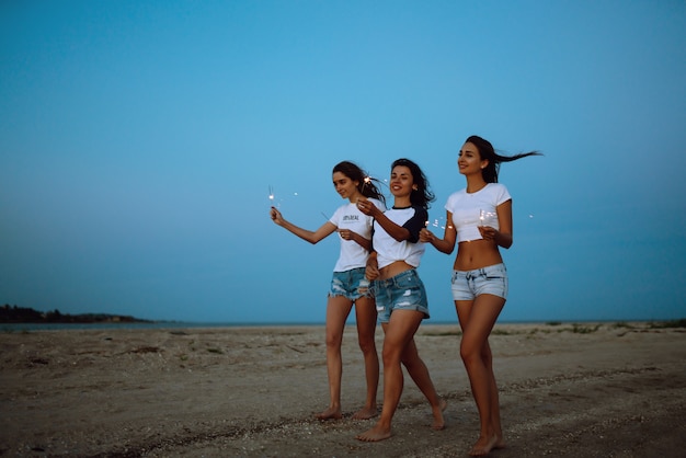 Os estrelinhas nas mãos de jovens na praia. Três meninas curtindo a festa na praia com acendedor. Férias de verão, férias, relaxar e conceito de estilo de vida.