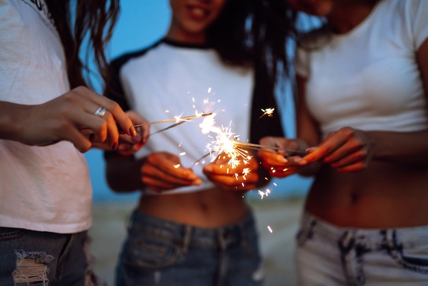 Os estrelinhas nas mãos de jovens na praia. Três meninas curtindo a festa na praia com acendedor. Férias de verão, férias, relaxar e conceito de estilo de vida.