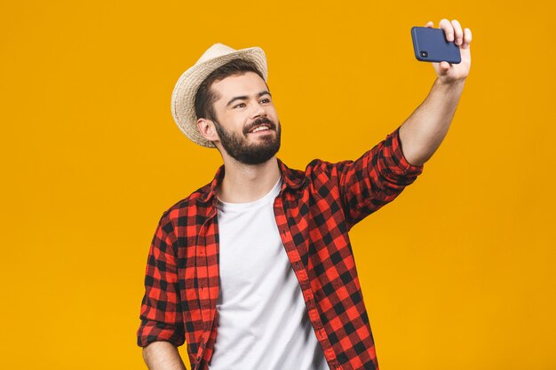 Os jovens alegres viajam o homem no chapéu que toma o selfie com o smartphone isolado sobre a parede amarela.