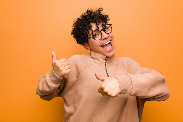 Os jovens misturaram a mulher afro-americano contra uma parede marrom que levanta ambos os polegares acima, sorrindo e confiantes.