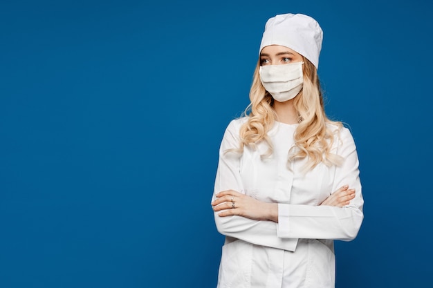 Os jovens nutrem em um vestido médico branco e em uma máscara médica em um azul, isolado. Médica em uniforme médico e máscara facial. Clínico geral ou estagiário posando em uniforme branco, cuidados de saúde.