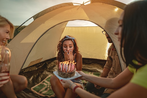 Os jovens se divertem no acampamento na natureza. Eles estão comemorando um aniversário, rindo e cumprimentando seu amigo com bolo de aniversário, felizes por estarem juntos.