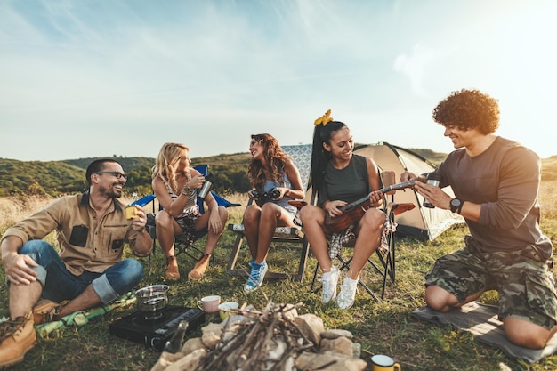 Os jovens se divertem no acampamento na natureza. eles estão descansando, rindo e cantando ao som de ukulele, felizes por estarem juntos.