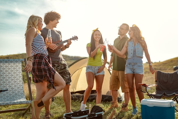 Os jovens se divertem no acampamento na natureza. Eles estão descansando, rindo e cantando com música de ukulele, felizes por estarem juntos.