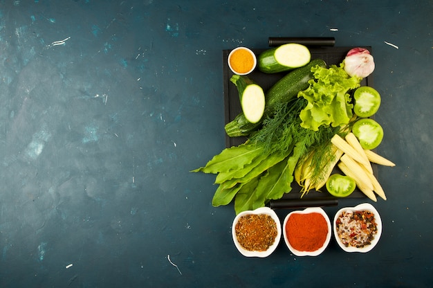 OS LEGUMES NA CAIXA NA PEDRA UM ESCURO. JOVENS VERDES CEBOLA ALHO ESPECIARIAS BRILHANTES COLOCAM-SE EM UMA BANDEJA DE MADEIRA COM PUNHOS EM UM TEXTURIZ ESCURO.