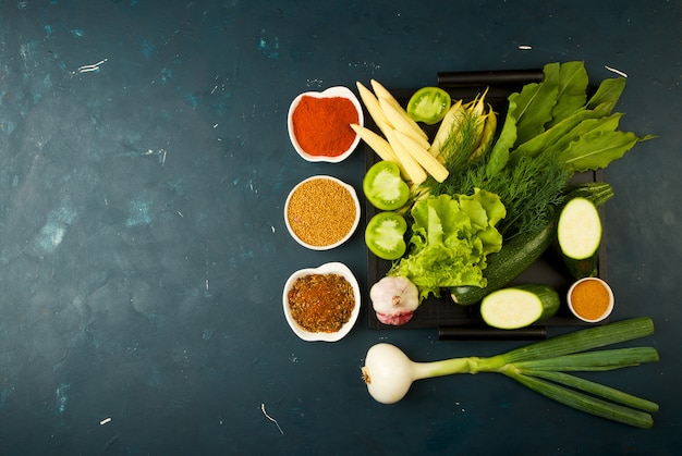 OS LEGUMES NA CAIXA NA PEDRA UM ESCURO. JOVENS VERDES CEBOLA ALHO ESPECIARIAS BRILHANTES COLOCAM-SE EM UMA BANDEJA DE MADEIRA COM PUNHOS EM UM TEXTURIZ ESCURO.