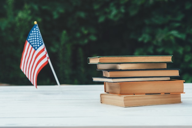 Os livros estão em uma mesa branca, no fundo é uma bandeira americana e folhas verdes.