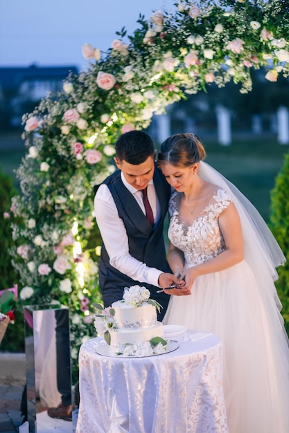 Os noivos estão cortando o bolo de casamento Os noivos no casamento cortam um lindo bolo branco decorado com flores