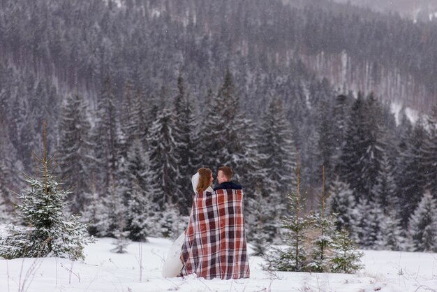 Os noivos ficam de pé contra o pano de fundo das montanhas e se aconchegam sob um tapete para se aquecer. Casamento de inverno