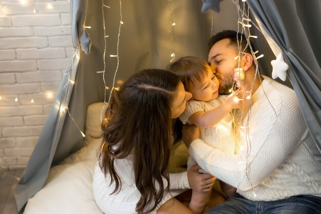 Os pais beijam a filha sentados sob uma guirlanda de luzes de Natal