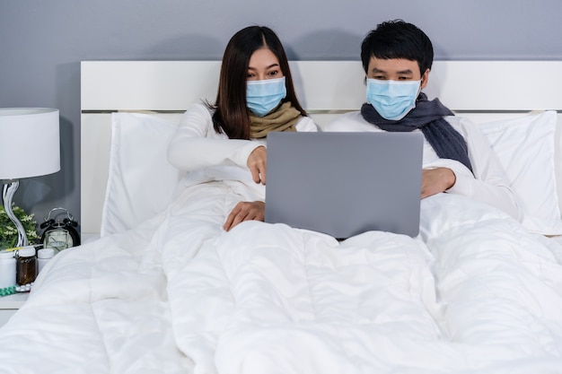 Os pares com máscaras médicas usando o laptop na cama durante o coronavírus pandêmico de quarentena ficam em casa.