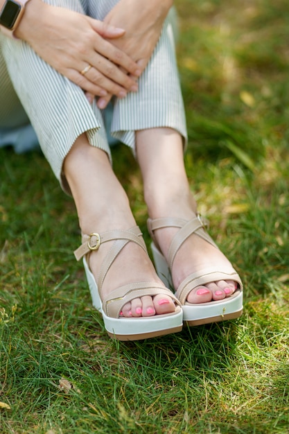 Os pés de uma mulher em sapatos de verão e calças despojadas em pé na grama verde