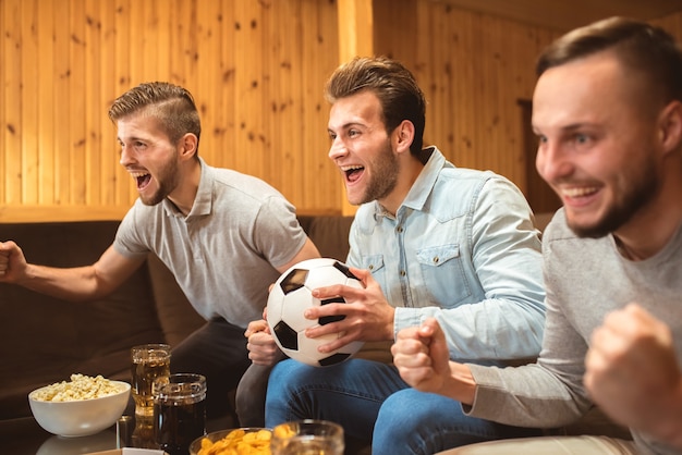 Foto os três homens com uma cerveja e uma comida assistem a uma bola de futebol e gesticulam