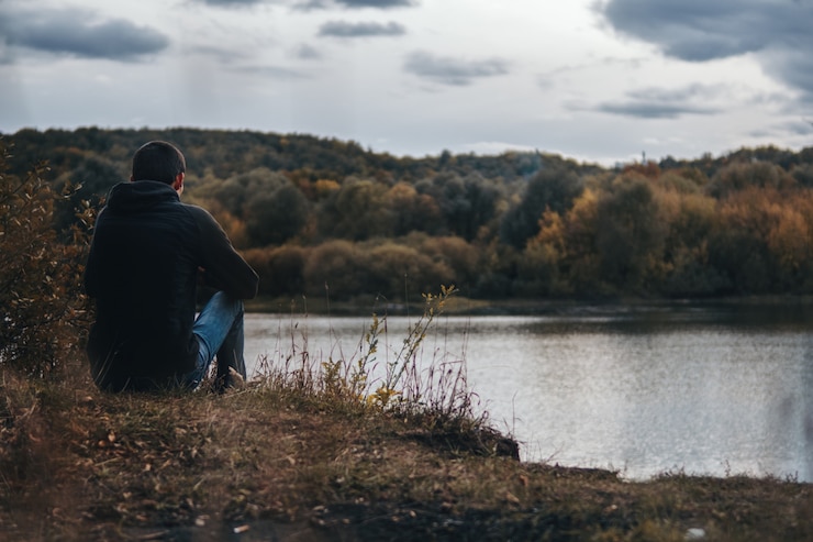 Foto oscuro otoño nublado. el chico sentado en la orilla del río desde atrás. soledad