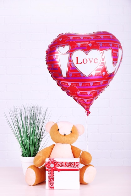 Oso de peluche con caja de regalo, planta y globo de corazón de amor en la mesa de madera, en el fondo de la pared de ladrillo