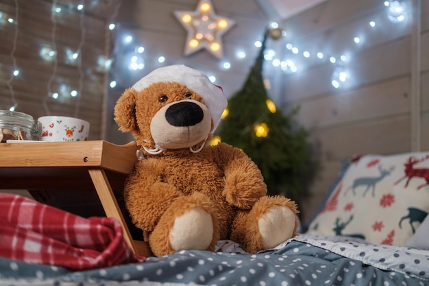 Un oso de peluche de juguete con un gorro de Papá Noel se sienta en la cama con el telón de fondo de un árbol de Navidad y una guirnalda brillante