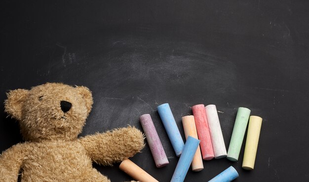 Oso de peluche marrón y crayones de colores sobre pizarra negra, regreso a la escuela