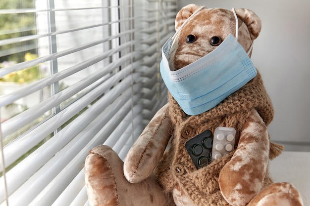 Un oso de peluche con una máscara médica mira con tristeza por la ventana. Niño enfermo en cuarentena