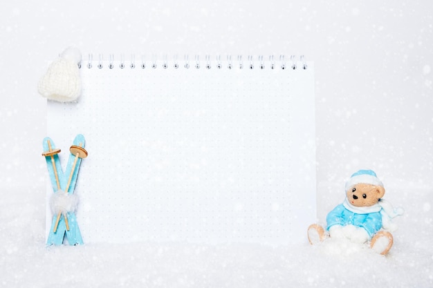Oso de peluche con ropa azul sentado en un trineo, esquís de madera azul, un sombrero blanco y un cuaderno en blanco sobre un fondo de nieve blanca y copos de nieve. Concepto de lista de tareas pendientes.