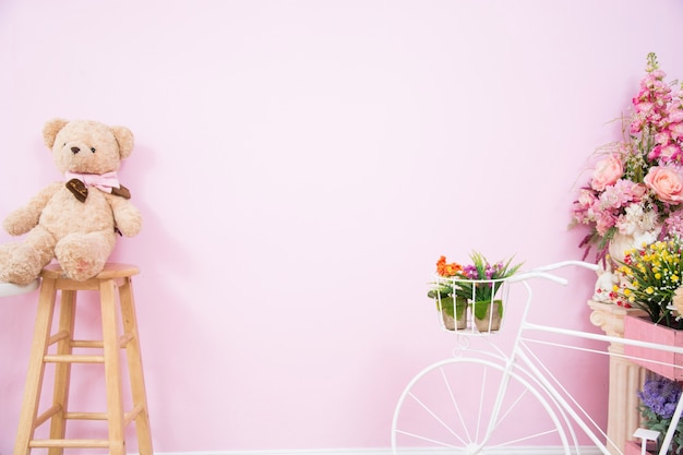 Oso de peluche en una silla de madera con flores y una bicicleta blanca con paredes de color rosa.