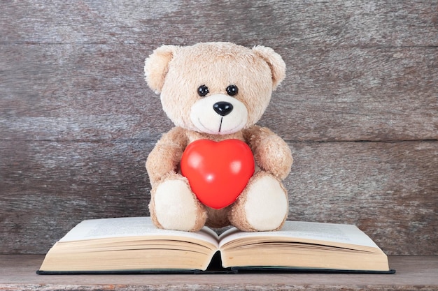 Oso de peluche sosteniendo un corazón rojo sentado en un libro antiguo y conceptos de fondo de madera de educación, amor y salud