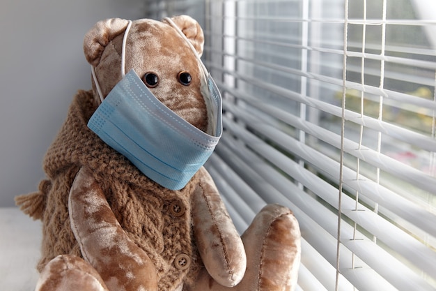 Un oso de peluche triste con una máscara médica mira por la ventana