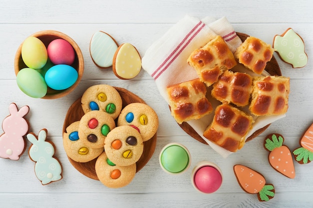 Oster-Hot-Cross-Brötchen mit farbigen Eiern Oster-Lebkuchen-Kaninchen und Kekse auf weißem Plankenholz-alten Tischhintergrund Frühlingsferien-Backkonzept Ostern-Tischeinstellung Draufsicht Mock-up