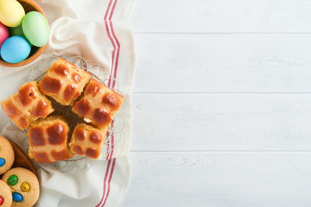 Oster-Hot-Cross-Brötchen mit farbigen Eiern Oster-Lebkuchen-Kaninchen und Kekse auf weißem Plankenholz-alten Tischhintergrund Frühlingsferien-Backkonzept Ostern-Tischeinstellung Draufsicht Mock-up