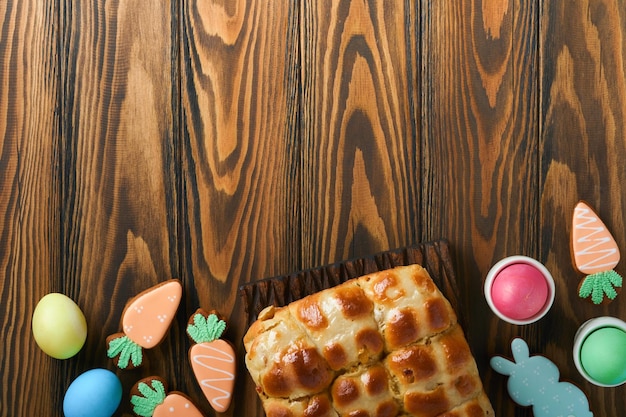 Oster-Hot-Cross-Brötchen mit farbigen Eiern Oster-Lebkuchen-Kaninchen und -Plätzchen über dunkelbraunem Brett aus Holz, alter Tischhintergrund Frühlingsferien-Backkonzept Ostern-Tischeinstellung Draufsicht Mock-up