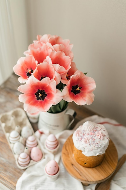 Oster-Panettone und gefärbte Ostereier. Ostersymbole. Rosa Tulpen im Metallkrug auf Holztisch