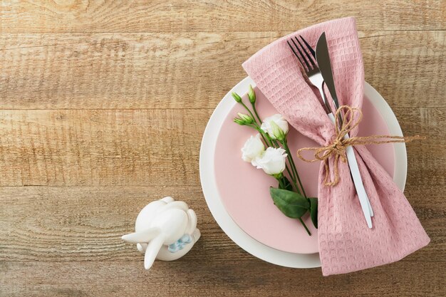 Osterbrunch Tischdekoration Frühlings-Osterferienkonzept mit weißem rosa Teller und Serviette mit dekorativen Symbolen Feiertagskanincheneier und Blumenstrauß auf altem hölzernem Hintergrund Draufsicht