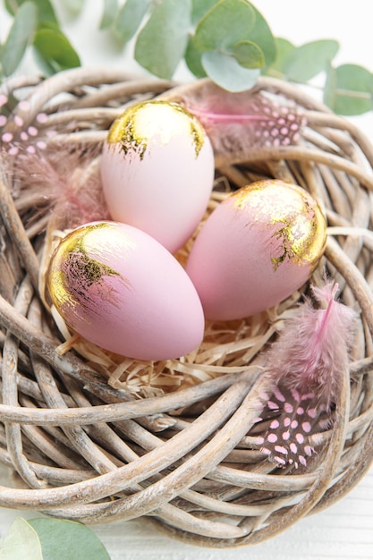 Ostereier gemalt in Rosa und Gold und Federn in einem Nest auf weißem Holzhintergrund