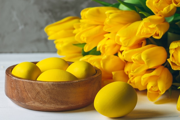 Ostereier in der Holzschale und im Strauß der leuchtend gelben Tulpen auf weißer Holzoberfläche auf grauer Oberfläche