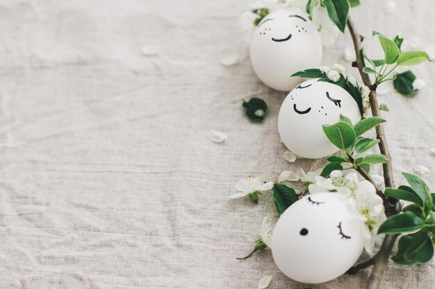 Ostereier mit gezeichneten lustigen Gesichtern in niedlichen Blumenkränzen auf Leinenstoff mit blühendem Zweig