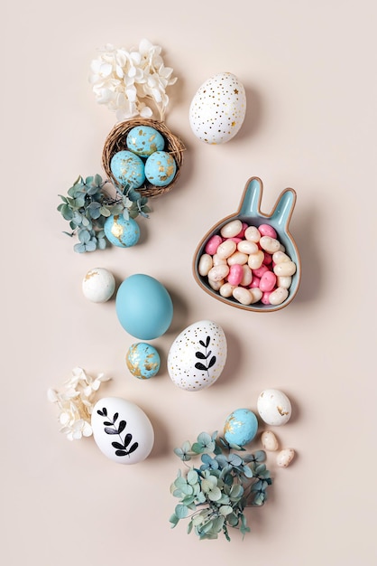 Ostereier mit Süßigkeiten und Blumen auf beige Fröhliches Osterkonzept Weiße und blaue Eier mit süßem Teller in Form eines Häschens