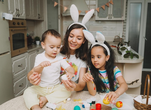 Osterfamilientraditionen Liebevolle junge Mutter, die Kindern beibringt, wie man Eier für Ostern malt, während sie zusammen am Küchentisch sitzt