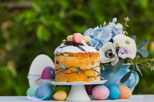 Osterferienkonzept Orthodoxes süßes Brot kulich mit Zuckerguss und Macarons auf der Oberseite Blumen in blauer Vase bunt bemalte Eier Wunderschöne traditionelle Osterszene im Frühling