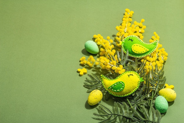 Osterkomposition mit traditionellen Frühlingsblumen und handgefertigten Filzvögeln. Dekorative Eier, harter, heller, dunkler Schatten, savannengrüner Farbhintergrund, Kopierraum