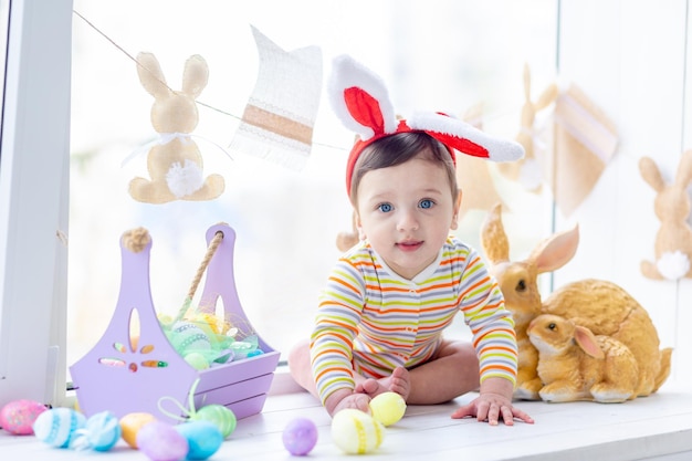 Osterkonzept Ein fröhliches Baby mit Hasenohren sitzt mit einem Kaninchen und Ostereiern am Fenster