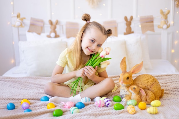 Ostern ein kleines Mädchen mit bemalten farbigen Eiern und ein Kaninchen, das Frühlingsblumen-Tulpen zu Hause in einem hellen Raum hält, der sich auf den Urlaub vorbereitet, lächelt und Spaß hat