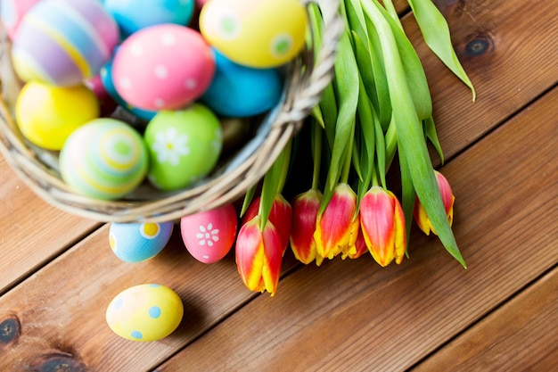 ostern, feiertage, tradition und objektkonzept - nahaufnahme von farbigen ostereiern im korb und tulpenblumen auf holztisch mit kopierraum