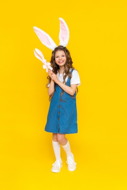 Ostern für Kinder Glückliches Baby mit langen Hasenohren Der Frühlingsurlaub ist Ostern Ein schönes kleines Mädchen mit langen Haaren in einem blauen Sommerkleid in voller Länge auf einem gelb isolierten Hintergrund